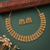 An ornate gold-toned traditional necklace with matching earrings, displayed on a dark green background with decorative dried flowers and a carved wooden prop.