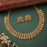 An ornate gold-toned traditional necklace with matching earrings arranged on a dark green surface, with a carved wooden prop at the top and small dried flowers at the lower left.