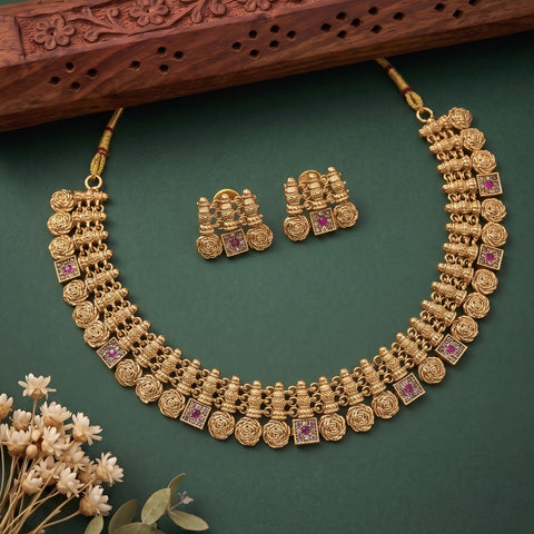An ornate gold-toned traditional necklace with matching earrings arranged on a dark green surface, with a carved wooden prop at the top and small dried flowers at the lower left.