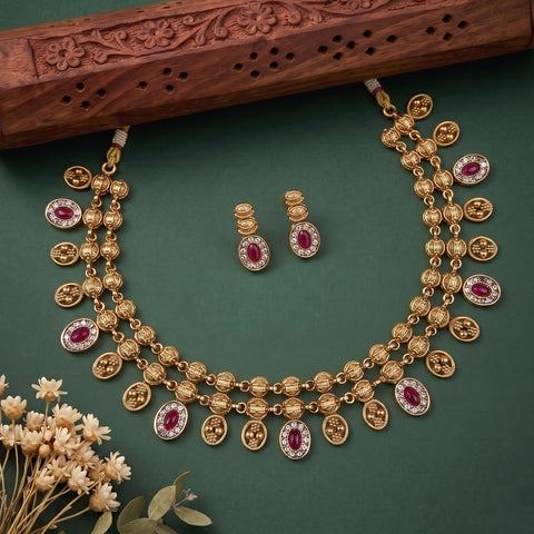Gold-toned traditional necklace and matching earrings with oval red gemstones and small clear stones, arranged on a dark green background with a carved wooden prop and dried flowers.