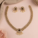 Gold-toned traditional jewelry set featuring a necklace and matching earrings with pearl-like drops and dark oval center stones, arranged on a soft pink background with neutral fabric props.