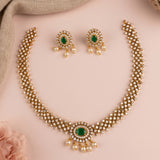 Gold-plated Indian ethnic jewelry set featuring a multi-strand necklace with a central green stone and pearl drops, matching drop earrings, and a beige fabric backdrop on a soft pink background.