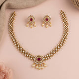 Gold-plated Indian traditional jewelry set featuring a necklace and matching earrings with white faux pearls and red oval gemstones, arranged on a soft pink background.
