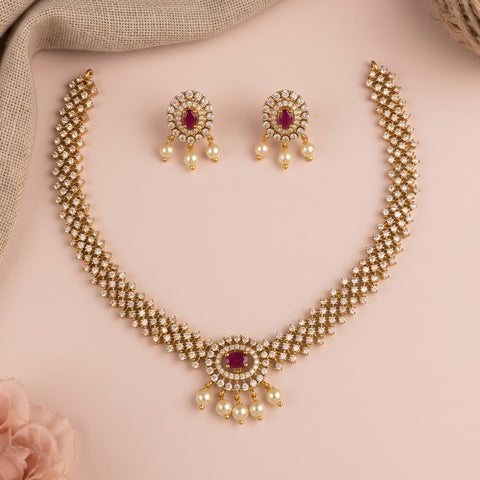 Gold-plated Indian traditional jewelry set featuring a necklace and matching earrings with white faux pearls and red oval gemstones, arranged on a soft pink background.