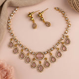Gold-tone statement necklace and matching drop earrings set with pale pink teardrop gemstones and clear rhinestone accents, arranged on a soft pink background with decorative fabric and a flower.