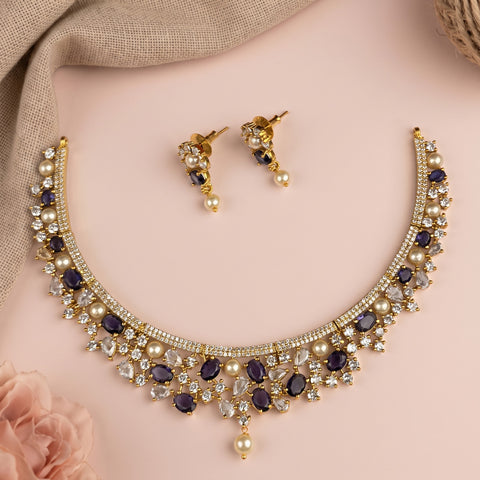 Gold-tone jewelry set featuring a curved necklace and matching drop earrings, decorated with dark purple oval stones, clear rhinestones, and white faux pearls, arranged on a soft pink background with a beige fabric drape.