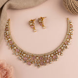 Gold-plated Indian-style bridal jewelry set featuring a choker necklace and matching drop earrings, embellished with light pink stones, clear crystals, and faux pearls, displayed on a soft pink background with decorative fabric and jute props.
