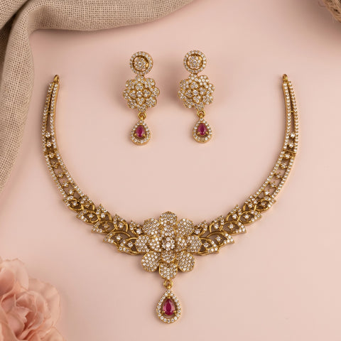 Gold-toned Indian bridal jewelry set featuring a choker necklace and matching drop earrings, embellished with clear pavé stones and red teardrop gemstones, displayed on a soft pink background.