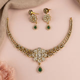 Gold-plated jewelry set featuring a necklace and matching earrings adorned with clear crystals and green teardrop gemstones, displayed on a soft pink background with a beige fabric accent.