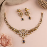 Gold-toned jewelry set with a necklace and matching earrings, decorated with clear rhinestones and dark blue teardrop stones, arranged on a soft pink background with decorative fabric and a rose.