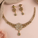 Gold-toned traditional jewelry set featuring a necklace and matching drop earrings, embellished with clear stones and light green teardrop accents, arranged on a soft pink background with a beige fabric corner.