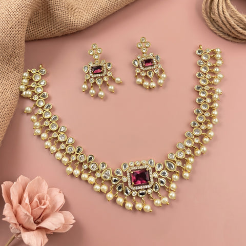 Gold-plated Indian Kundan jewelry set featuring a necklace, earrings, and a matching ring, decorated with clear crystals and pink gemstones, arranged on a dusty pink background with decorative props.