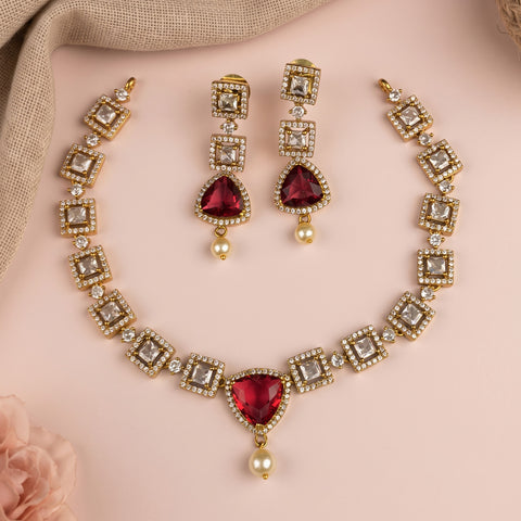 Gold-plated jewelry set featuring a necklace, earrings, and matching rings with clear rhinestones and deep red teardrop-shaped center stones, accented with small white pearls, arranged on a soft pink background.