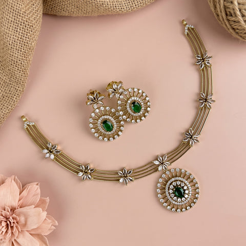 Gold-toned jewelry set with a necklace and matching earrings, featuring green oval stones and white crystal floral motifs, arranged on a soft pink background with burlap and a flower prop.
