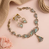 Gold-toned jewelry set with teardrop-shaped light green stones and crystal accents, arranged on a soft pink background with burlap, rope, and a pink flower prop.