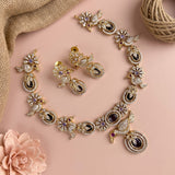 Gold-toned traditional jewelry set featuring a necklace, earrings, and bangles with intricate peacock motifs, embellished with clear rhinestones, black enamel, and purple gemstones, arranged on a soft pink background with burlap and a flower prop.