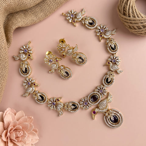 Gold-toned traditional jewelry set featuring a necklace, earrings, and bangles with intricate peacock motifs, embellished with clear rhinestones, black enamel, and purple gemstones, arranged on a soft pink background with burlap and a flower prop.