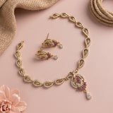 Gold-toned jewelry set with a matching necklace and earrings, featuring purple stone accents and clear crystal details, arranged on a soft pink background with burlap and a flower prop.