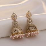 A pair of ornate gold-toned jhumka earrings with clear stones, pink bead drops, and white pearl accents, displayed on a soft cream satin fabric background.