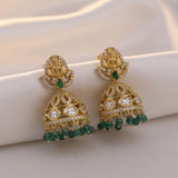 A pair of ornate gold-toned jhumka earrings with green stone accents and clear rhinestone details, displayed on a soft cream fabric background.
