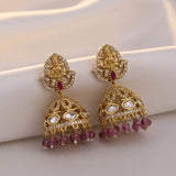 A pair of ornate gold-toned Indian jhumka earrings decorated with clear stones and small red gemstones, displayed on a draped cream satin fabric.
