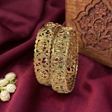 Two ornate gold-plated bangles with intricate filigree and red, green, and clear stone embellishments, displayed on a deep burgundy fabric backdrop next to white jasmine flowers and a carved wooden box.