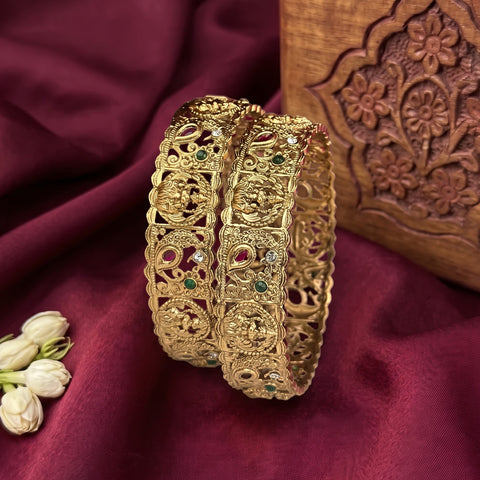 Two ornate gold-plated bangles with intricate filigree and red, green, and clear stone embellishments, displayed on a deep burgundy fabric backdrop next to white jasmine flowers and a carved wooden box.