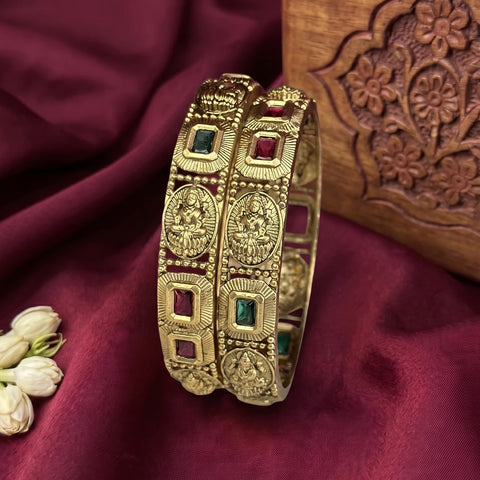 Two ornate gold-toned bangles with engraved deity motifs and small green and red stone accents, displayed on draped burgundy fabric with a carved wooden prop and white flowers.