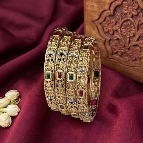 Four ornate gold bangles with intricate filigree work and small green and red stone accents, arranged on a deep burgundy fabric backdrop with a carved wooden block and white flower buds as props.