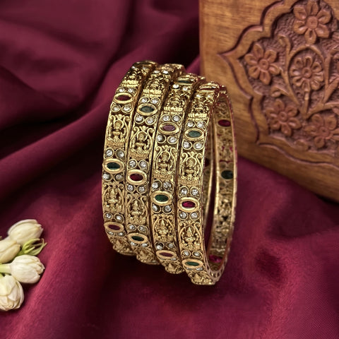 Four ornate gold bangles with intricate filigree work and small green, red, and white stone accents, displayed on a deep maroon satin fabric next to a carved wooden box.