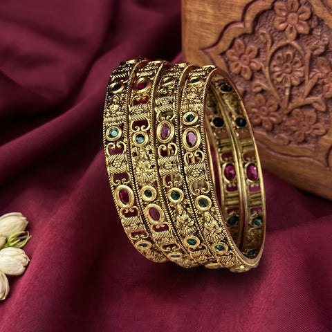 A set of five ornate gold-toned bangles decorated with red, green, and blue stones, arranged on a draped deep maroon fabric with a carved wooden prop.