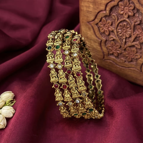 A gold-toned multi-strand bangle bracelet decorated with small Buddha motifs and red, green, and clear rhinestones, displayed on draped burgundy fabric next to a carved wooden box.
