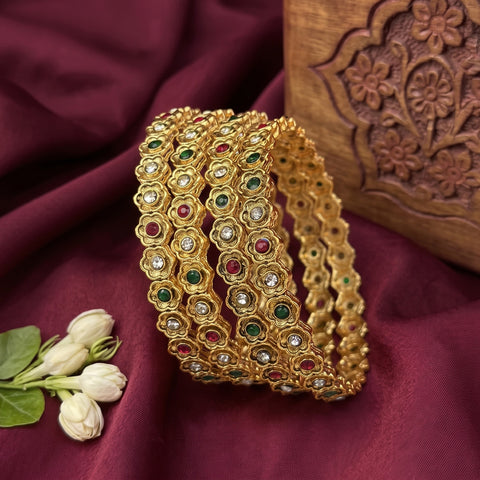 A gold-plated Indian style bangle bracelet with floral motifs and small red, green, and white stones, displayed on a deep maroon fabric next to white rose buds and a carved wooden box.