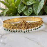 An ornate gold-toned traditional Indian headband (tilak) decorated with floral engravings, a central Ganesha motif, and dangling green bead chains, displayed on a white marble surface with green plants in the background.