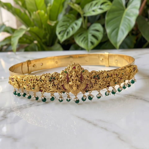 An ornate gold-toned traditional Indian headband (tilak) decorated with floral engravings, a central Ganesha motif, and dangling green bead chains, displayed on a white marble surface with green plants in the background.