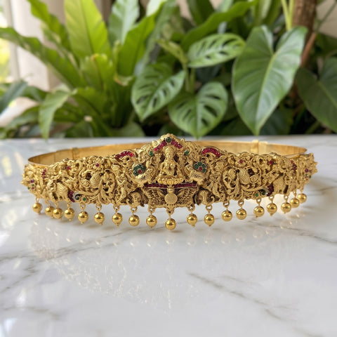 An ornate gold-toned traditional headpiece or crown with intricate filigree detailing, red and green stone accents, and dangling gold beads, displayed on a white marble surface with green plants in the background.