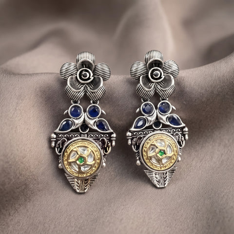 A pair of ornate silver-toned earrings with blue gemstone accents and a gold-toned circular center detail, displayed on draped taupe fabric.
