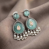 A pair of ornate dangle earrings with light green cat's eye stones, silver settings, and dangling pearl-like beads, displayed on a draped brown fabric background.