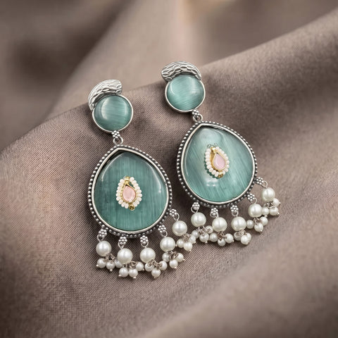 A pair of ornate dangle earrings with light green cat's eye stones, silver settings, and dangling pearl-like beads, displayed on a draped brown fabric background.