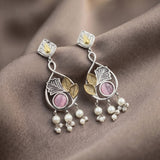 A pair of silver-toned dangle earrings with gold-toned leaf accents, pink oval stones, and dangling pearl-like beads, displayed on draped brown fabric.