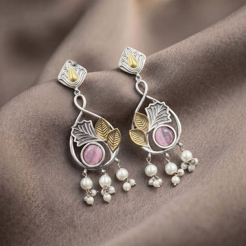 A pair of silver-toned dangle earrings with gold-toned leaf accents, pink oval stones, and dangling pearl-like beads, displayed on draped brown fabric.