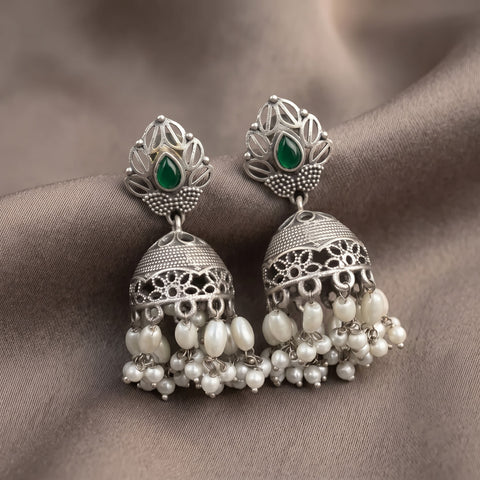 A pair of silver-toned Indian jhumka earrings with green teardrop stones and dangling white pearl-like beads, displayed on a draped taupe fabric background.