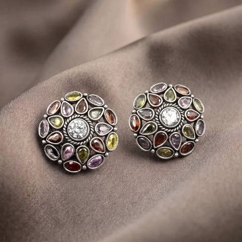 A pair of round silver-toned stud earrings decorated with multiple teardrop-shaped gemstones in assorted colors, including yellow, green, red, purple, and orange, arranged on a draped brown satin fabric.