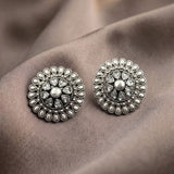 A pair of round silver-toned statement earrings with intricate floral detailing and clear teardrop gemstones, displayed on a draped taupe fabric background.
