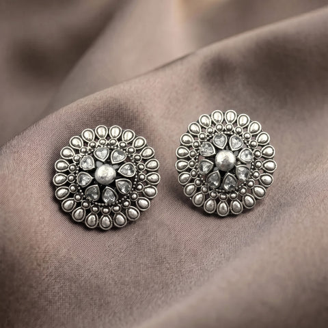 A pair of round silver-toned statement earrings with intricate floral detailing and clear teardrop gemstones, displayed on a draped taupe fabric background.