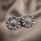 A pair of silver-toned floral stud earrings with small pinkish-red and clear stones, displayed on a draped taupe fabric background.