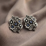 A pair of ornate silver-toned owl stud earrings with blue gemstone eyes and detailed feather patterns, displayed on draped brown fabric.