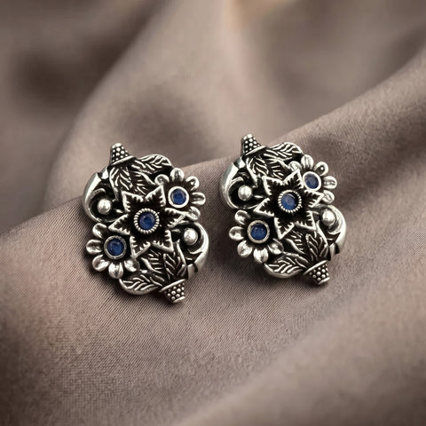 A pair of ornate silver-toned owl stud earrings with blue gemstone eyes and detailed feather patterns, displayed on draped brown fabric.