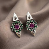 A pair of silver-toned geometric drop earrings with green round stones and a central purple stone, displayed on draped taupe fabric.