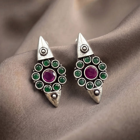 A pair of silver-toned geometric drop earrings with green round stones and a central purple stone, displayed on draped taupe fabric.